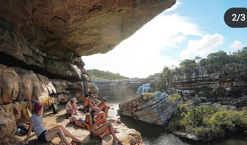 Hikers resting on rocks beneath a cave overhang watching Strandloper Falls cascade on the Wild Coast, South Africa