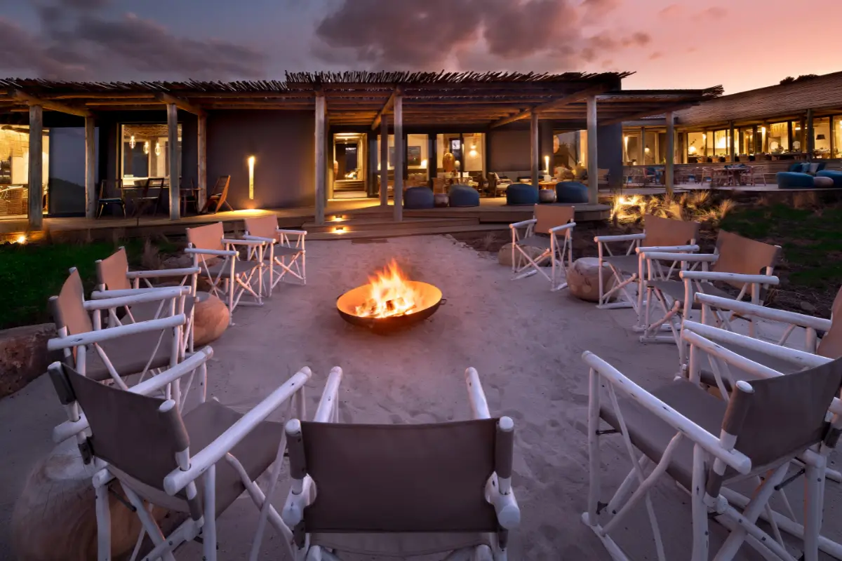 Outdoor fire pit ringed by canvas chairs at dusk at Gwegwe Beach Lodge, Mkambathi Wild Coast, with lodge lit up behind