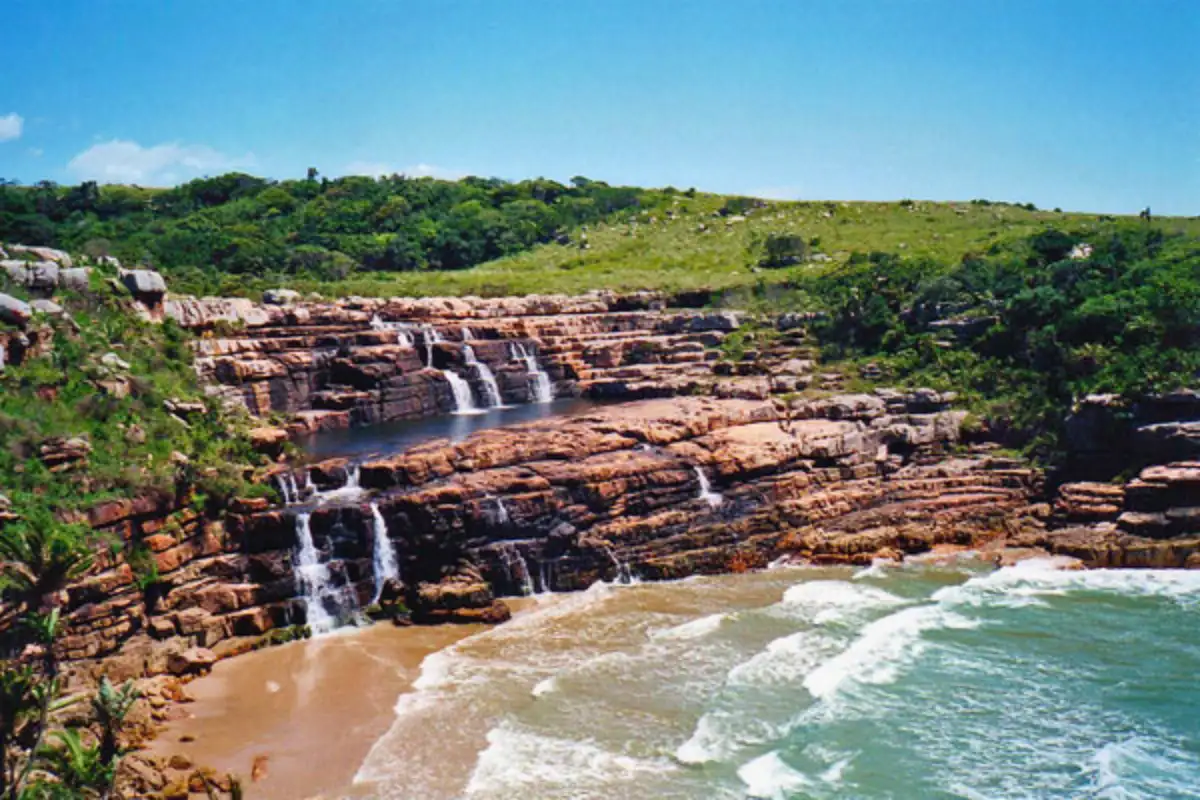Rocky waterfall cascading over red sandstone cliffs onto a Wild Coast beach near Mtentu, Eastern Cape South Africa